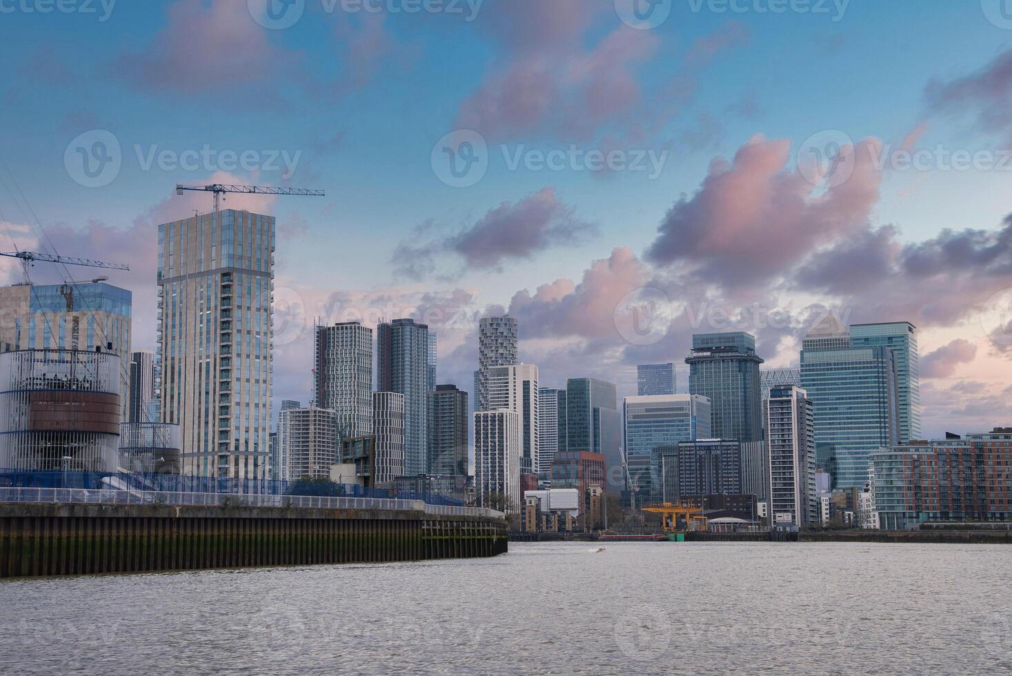 Modern skyscrapers of Canary Wharf in London with the River Thames in the foreground, construction cranes, and a partly cloudy early evening sky. photo