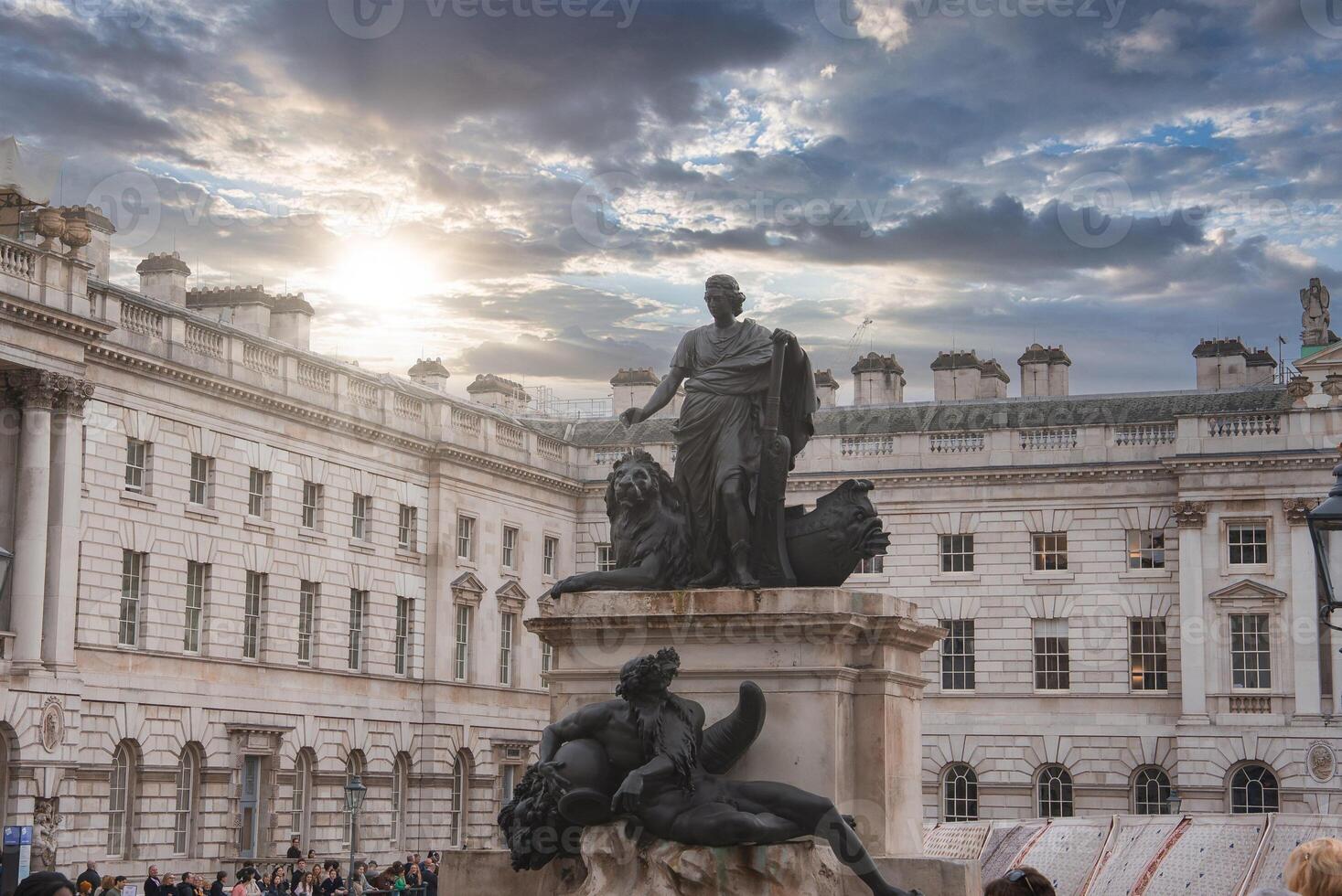 A statue with a lion and reclining figures stands in Somerset House courtyard, London, with neoclassical architecture and a dramatic sky in the background. photo