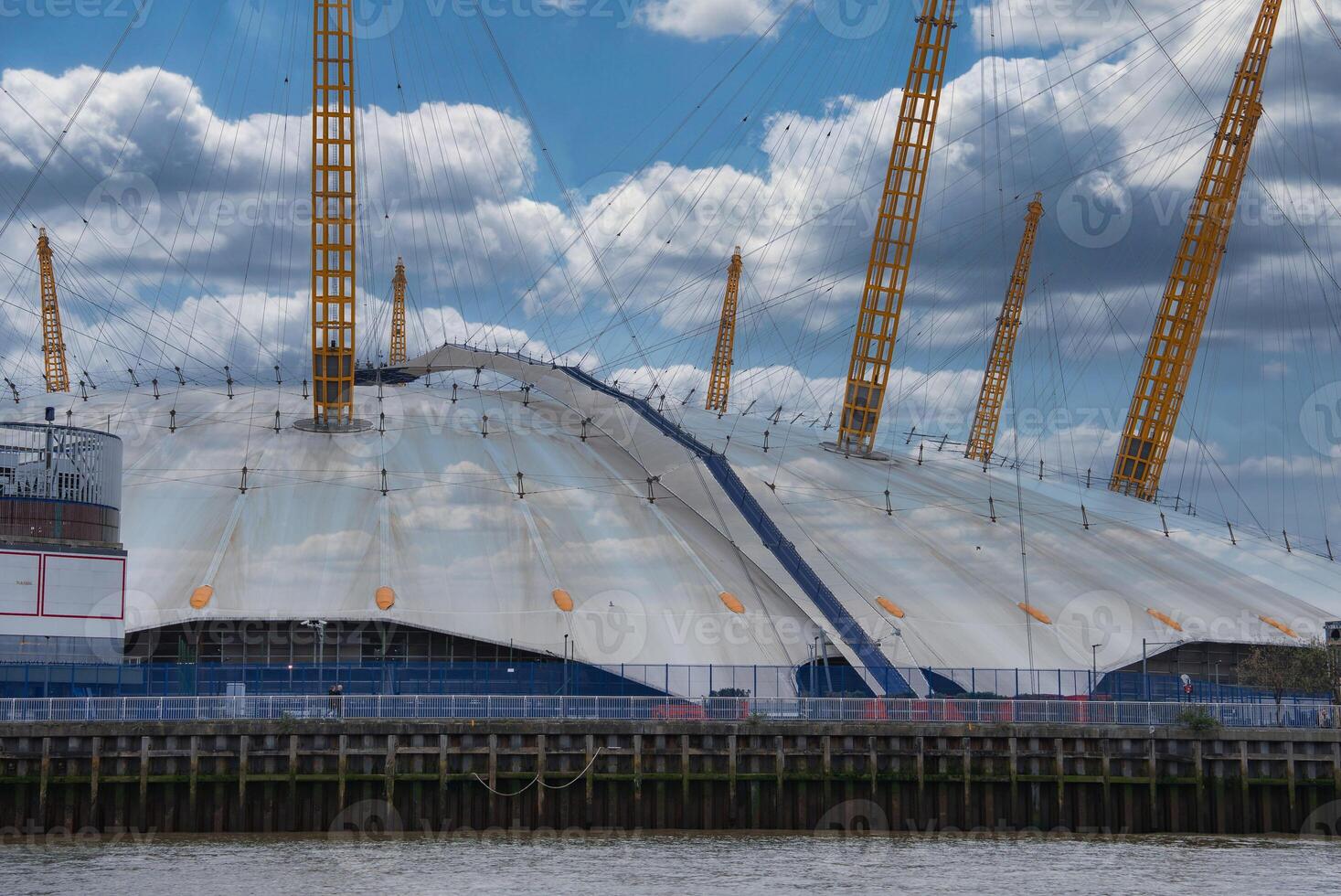 The O2 Arena in London, England, features its white dome and yellow masts. The River Thames flows in the foreground under a partly cloudy sky. photo