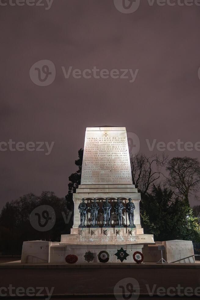 The Guards Memorial in London stands illuminated at night, with bronze soldier statues, detailed inscriptions, wreaths, and trees in the background. photo