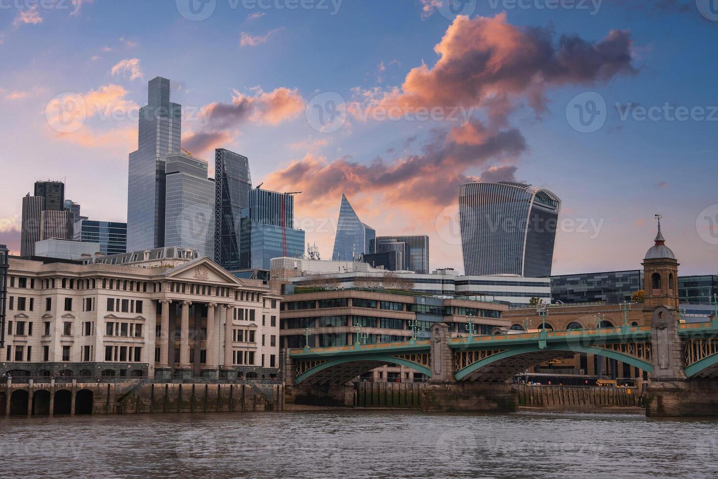 View of London's skyline featuring the Walkie Talkie and Gherkin buildings, Southwark Bridge over the River Thames, and a vibrant sunset sky. photo