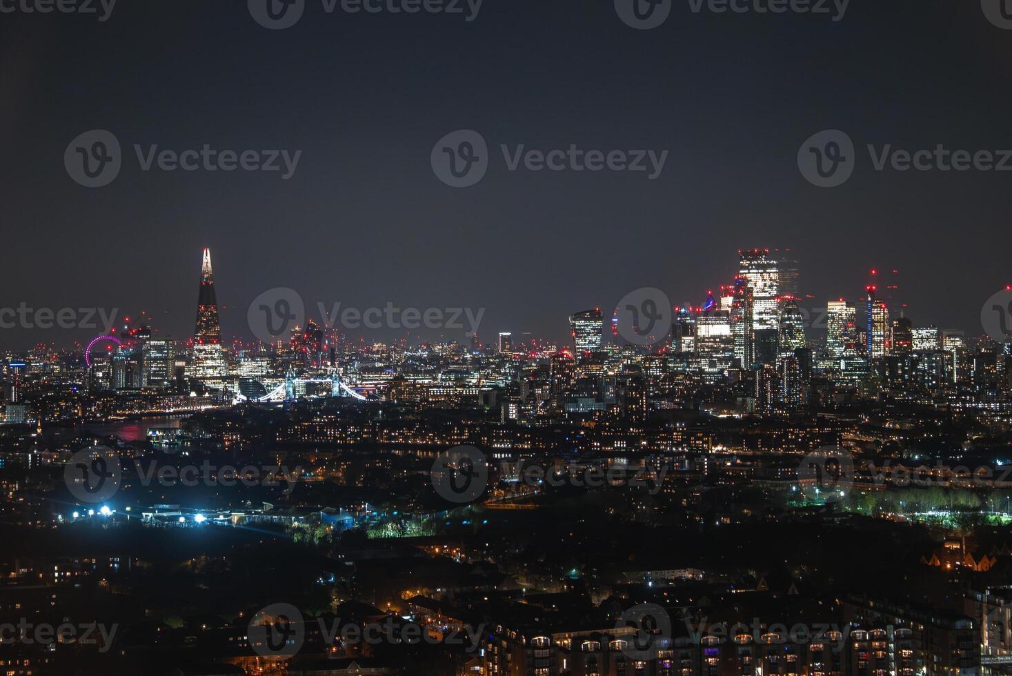 Illuminated London skyline at night with The Shard, Tower Bridge, modern skyscrapers, older architecture, residential areas, and greenery. photo