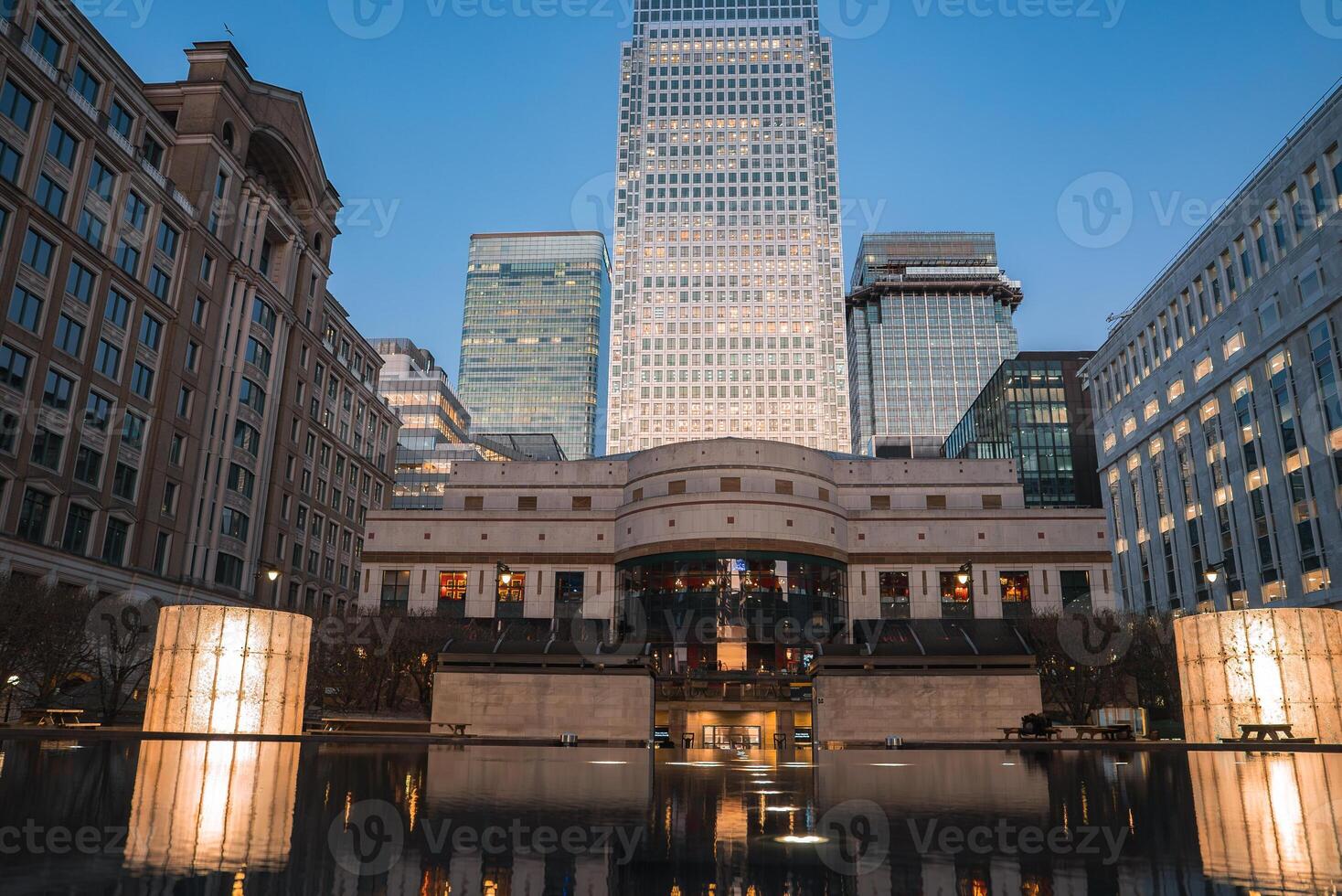 Twilight view of Canary Wharf in London featuring One Canada Square, modern office buildings, and a calm water feature reflecting the illuminated skyline. photo