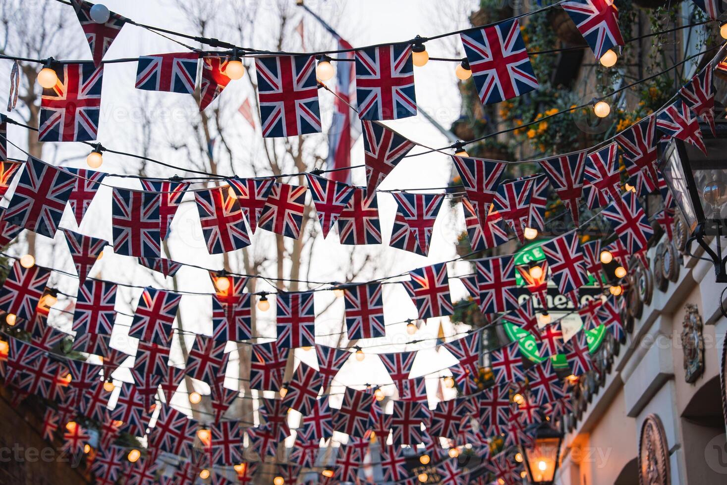 A London street decorated with Union Jack flags and warm string lights. Traditional architecture, bare trees, and greenery enhance the urban scene. photo