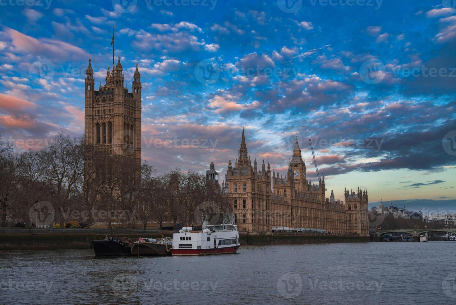 The Palace of Westminster with Gothic architecture stands by the River Thames at sunset. Boats are moored in the foreground under a partly cloudy sky. photo