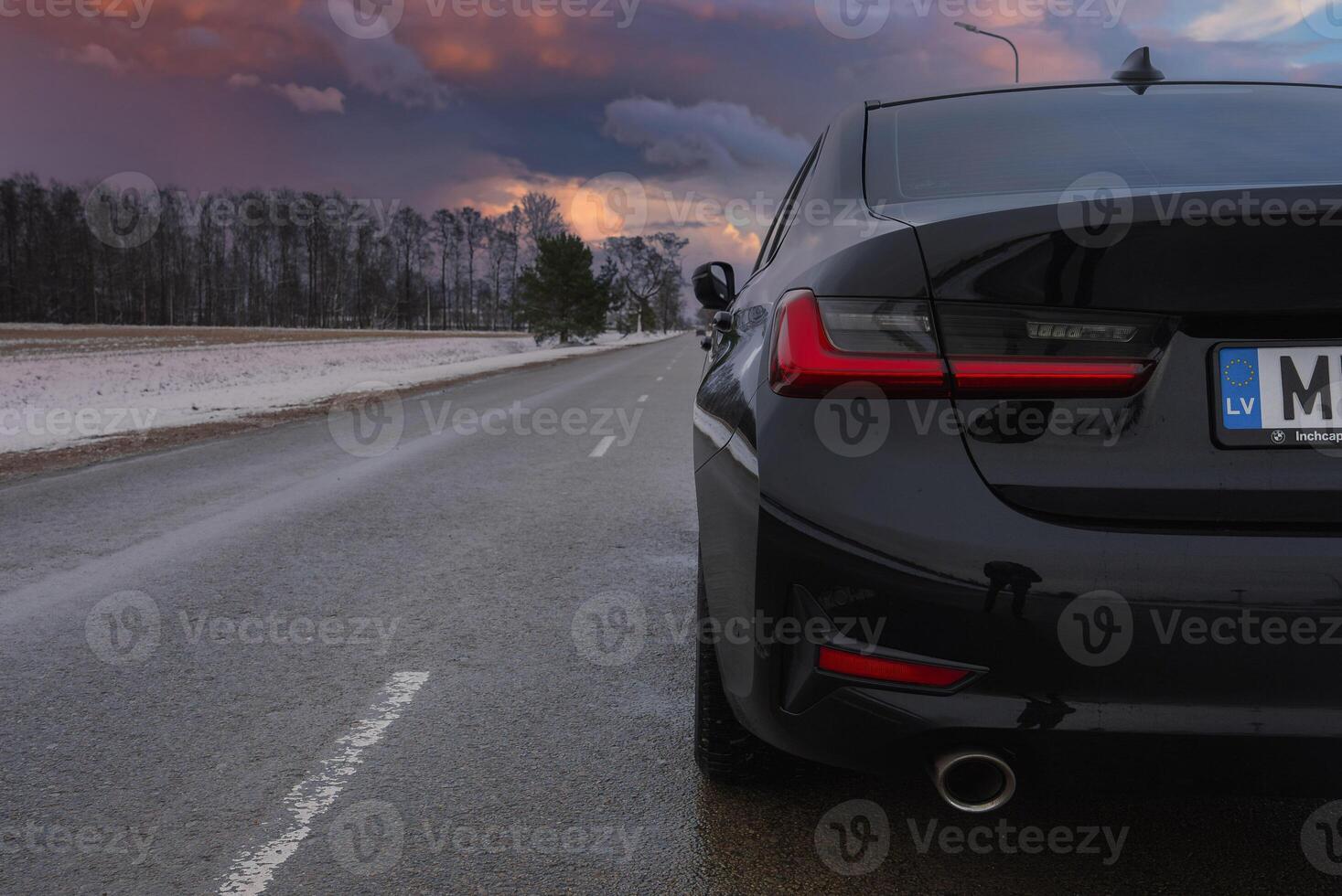 Rear view of a black BMW G20 320 with sharp taillights and exhaust pipe, parked on a wet road. Snowy roadside and bare trees under an overcast sky. photo