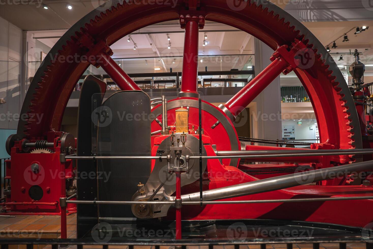 A red industrial steam engine with a massive flywheel and connecting rods, displayed indoors with protective railings in a modern London museum. photo