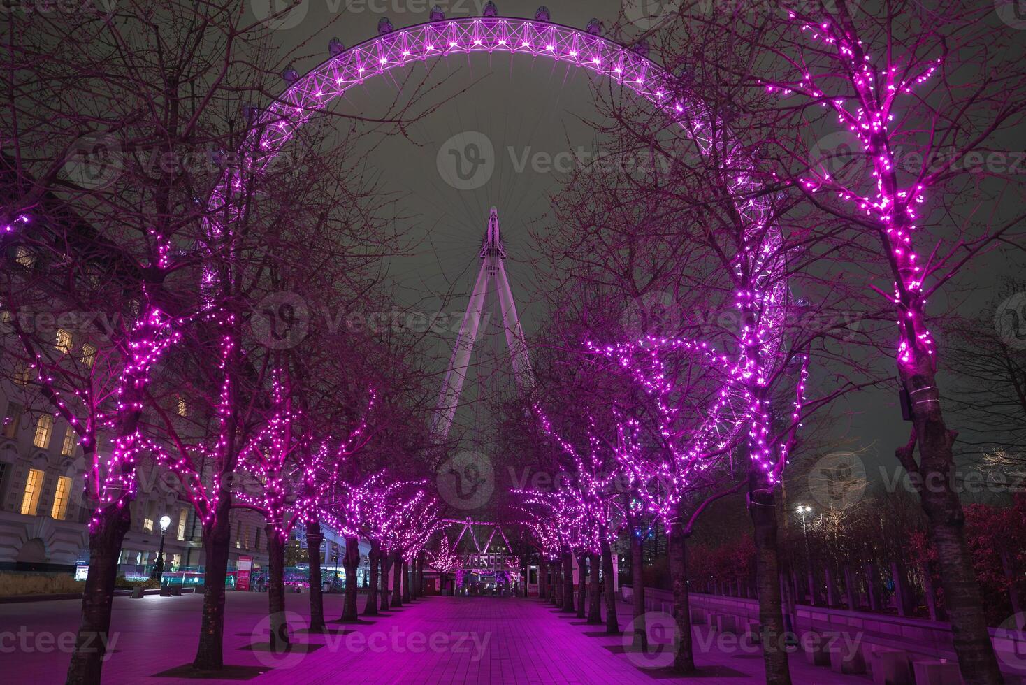 The London Eye is illuminated against the night sky, framed by a pathway of trees adorned with vibrant pink lights in a symmetrical arrangement. photo