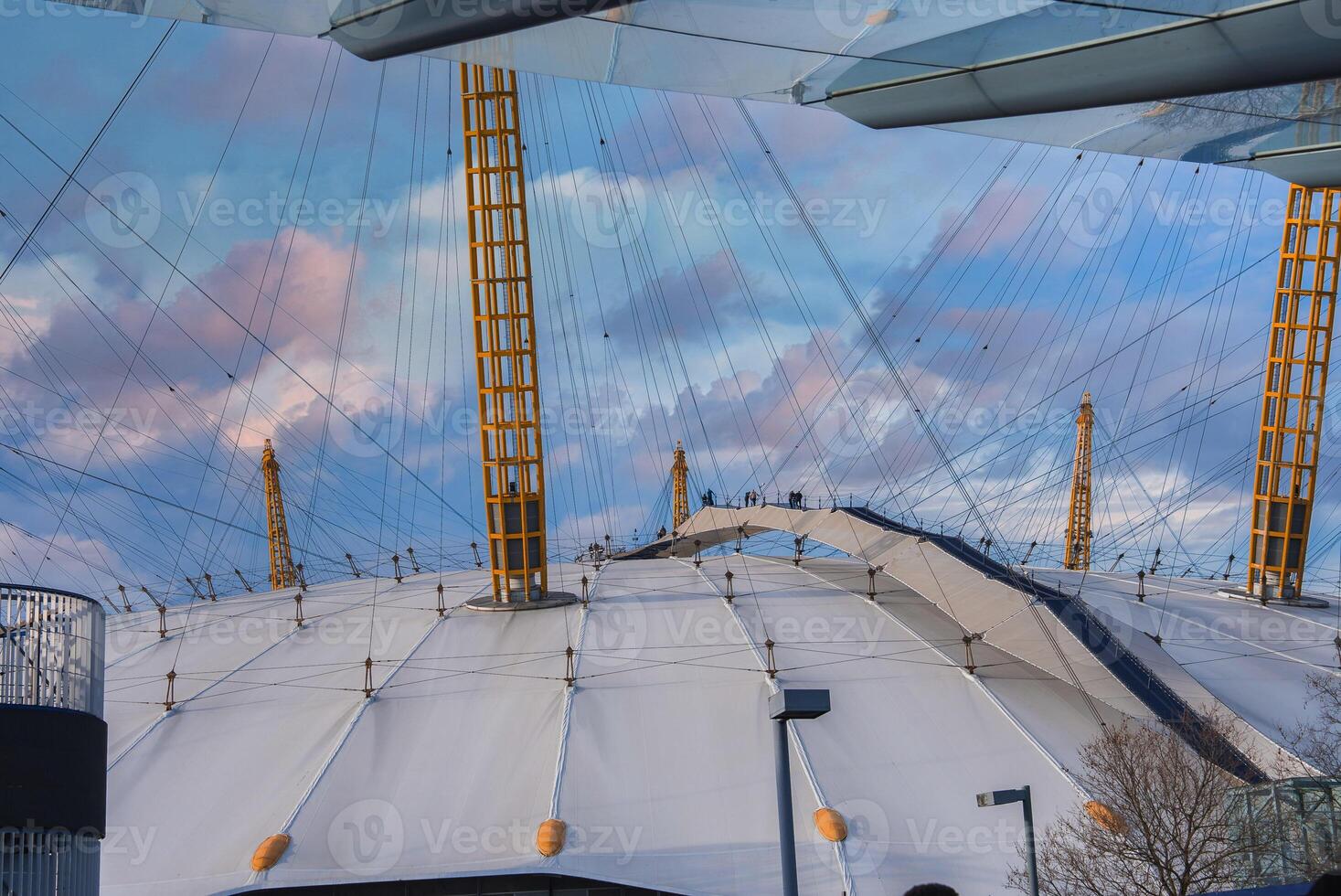 The O2 Arena in London features its white dome, yellow masts, and visible walkway. Modern buildings frame the scene under a blue and pink sky. photo