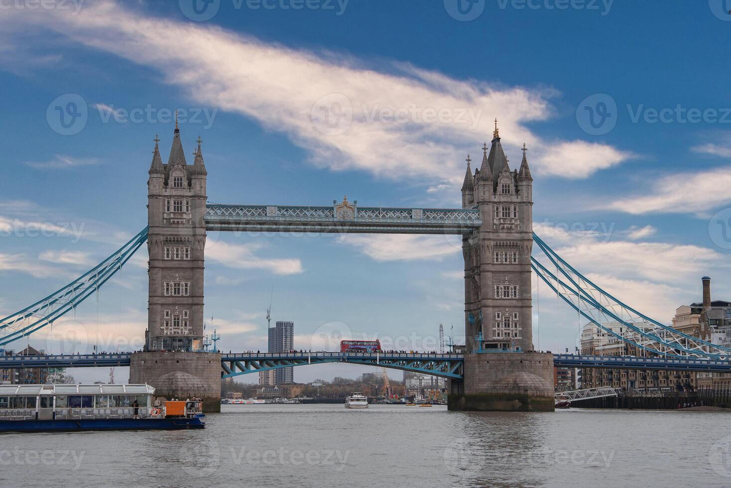 Tower Bridge in London, England, with its iconic towers and upper walkways, a red double decker bus crossing, and a docked boat in the foreground. photo
