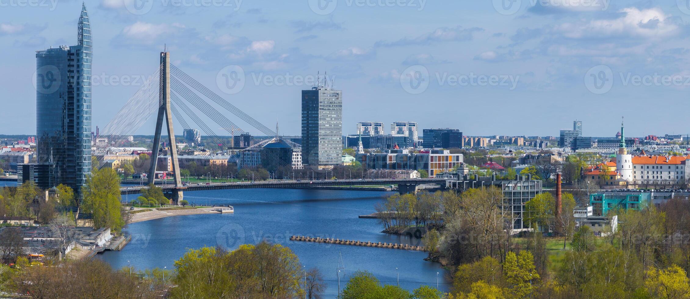 Aerial view of Riga, Latvia, showcasing the Vansu Bridge over the Daugava River, Z Towers, Riga Castle, lush greenery, and water channels under a partly cloudy sky. photo