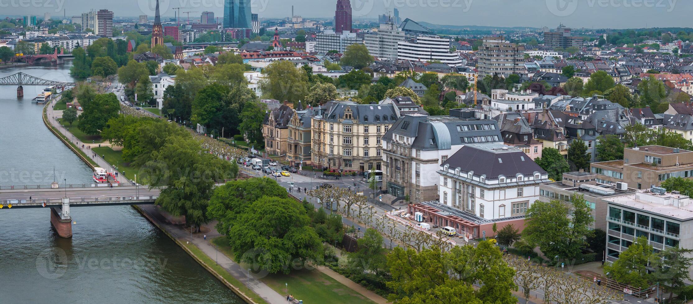 Aerial image of Frankfurt, Germany, featuring the Main River, a bridge, the European Central Bank, a Gothic church spire, and urban greenery. photo