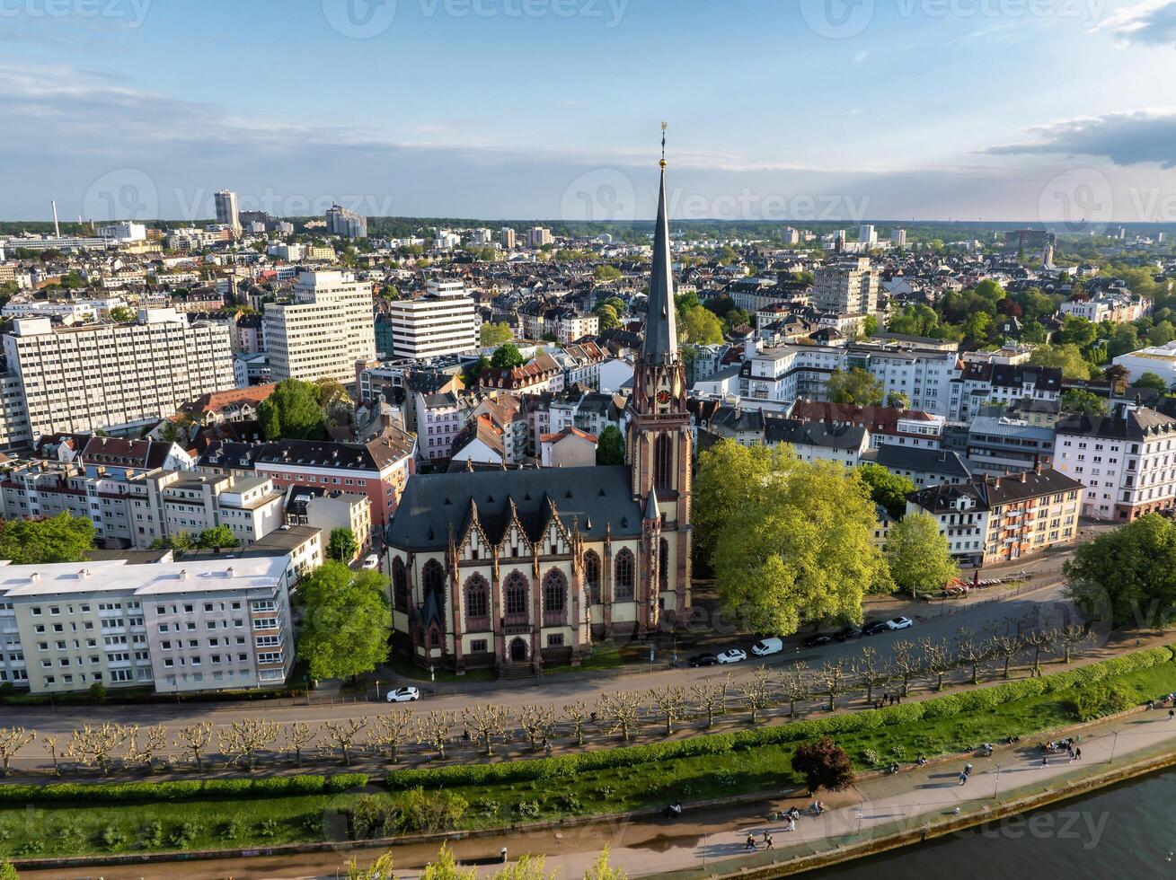 Aerial view of Frankfurt, Germany, highlighting Dreikonigskirche's Gothic spire, the Main River, tree lined pathways, and an expansive urban skyline. photo