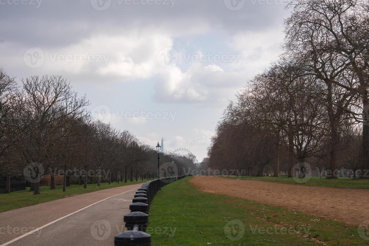A paved road and dirt path run parallel in a London park, separated by grass and lined with bare trees. The London Eye is visible under a partly cloudy sky. photo