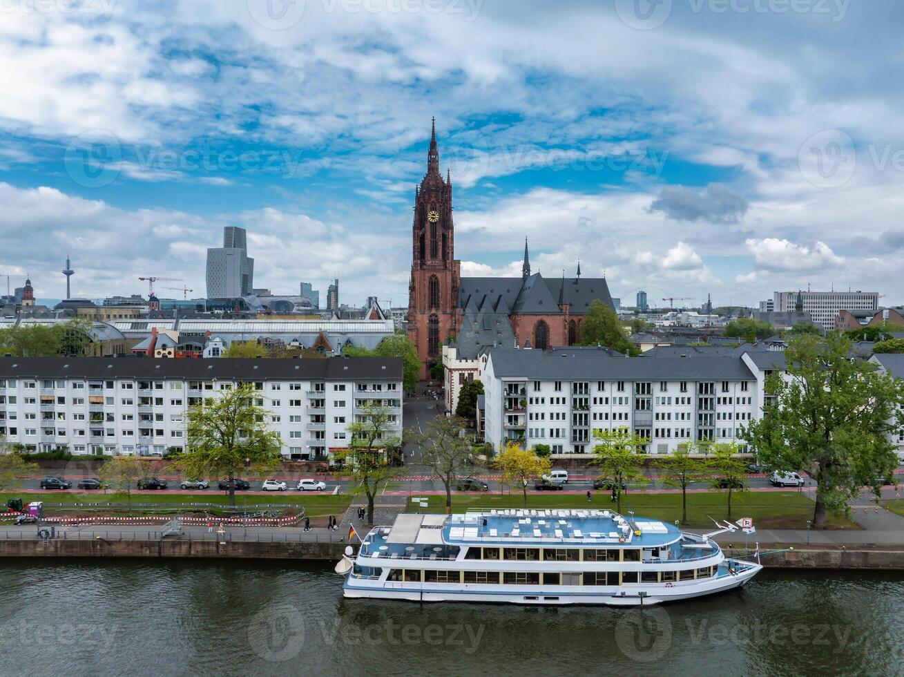 Aerial image of Frankfurt Cathedral, modern skyscrapers, and Europaturm. A river with a docked passenger boat is in the foreground under a partly cloudy sky. photo