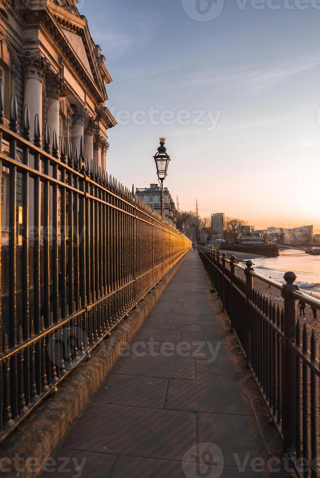 A riverside walkway in London during sunset, featuring a historic building, a traditional streetlamp, and the River Thames with modern buildings beyond. photo