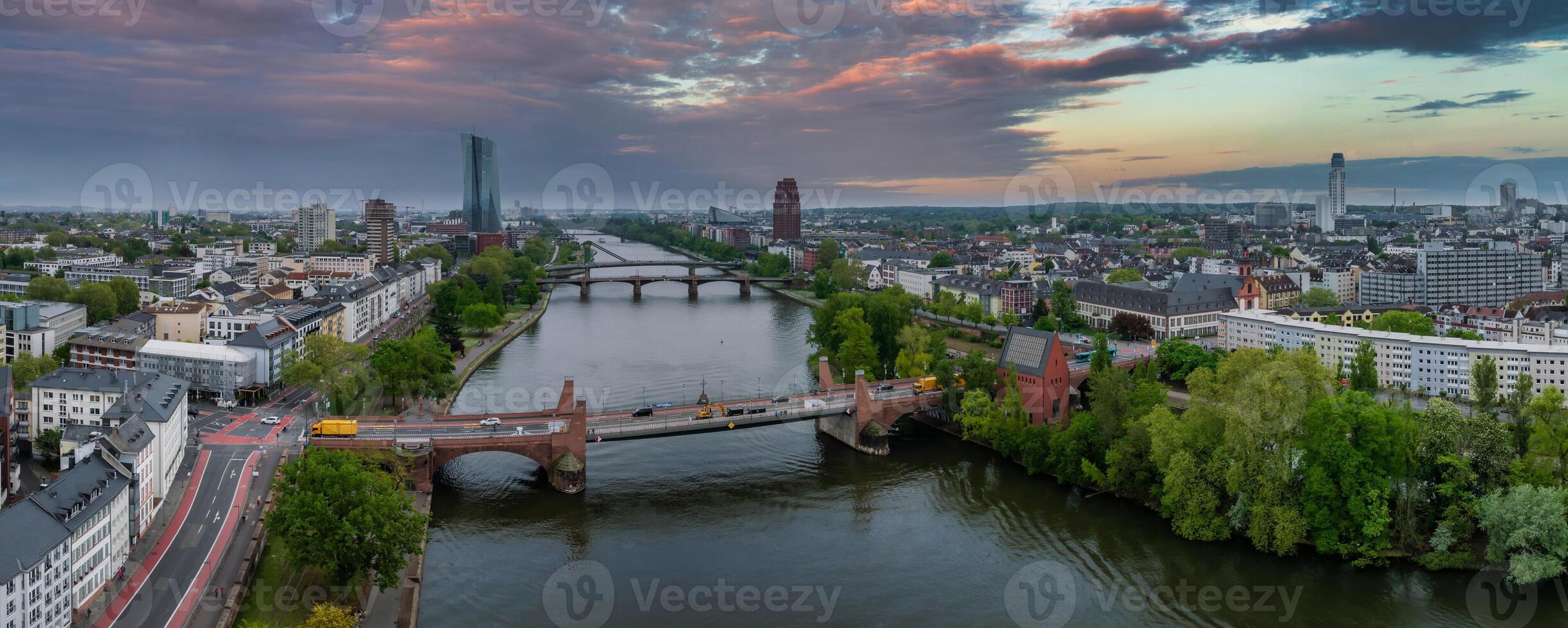Aerial view of Frankfurt, Germany, featuring the Main River, European Central Bank, bridges, lush greenery, and a dramatic orange and purple sunset. photo