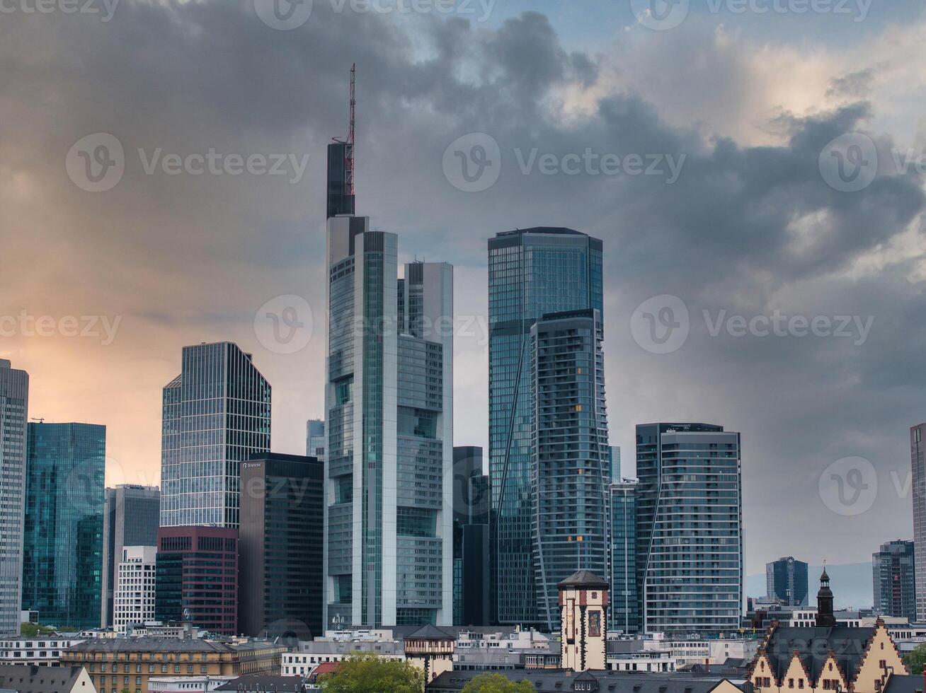 Frankfurt skyline at sunset featuring Commerzbank Tower, modern skyscrapers, a church with a clock tower, and dramatic clouds in the sky. photo