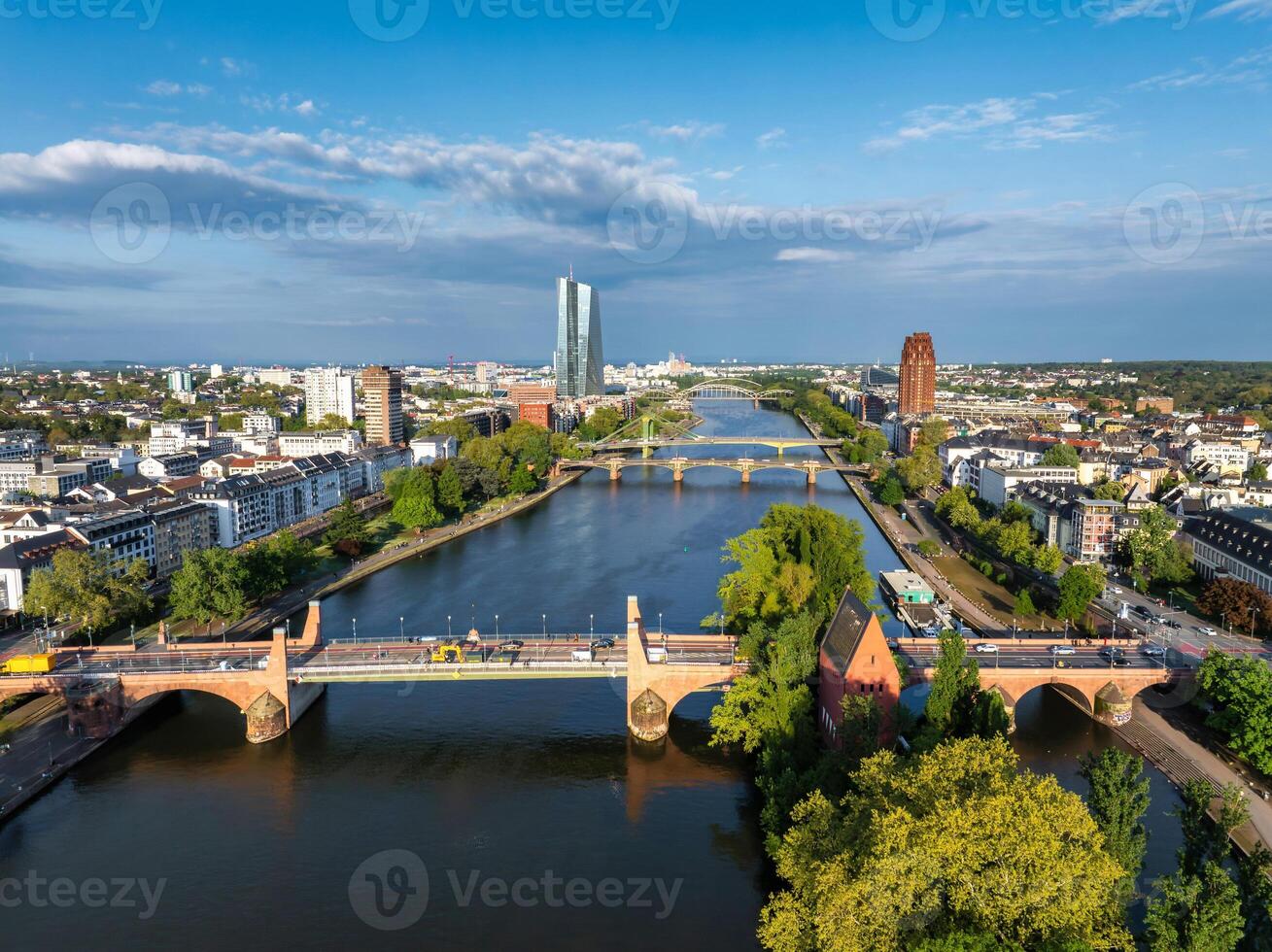 Aerial image of Frankfurt, Germany, featuring the Main River, European Central Bank, Alte Brucke, multiple bridges, and greenery under a clear blue sky. photo