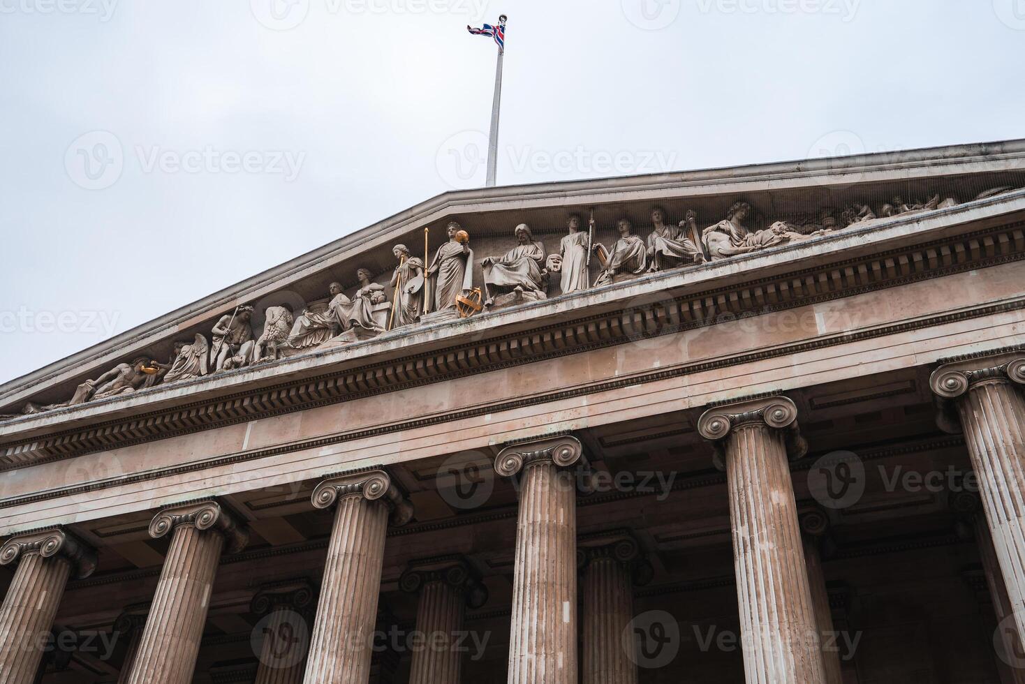 The facade of a neoclassical building in London features a triangular pediment with sculptures, fluted columns, and a Union Jack flag under a cloudy sky. photo