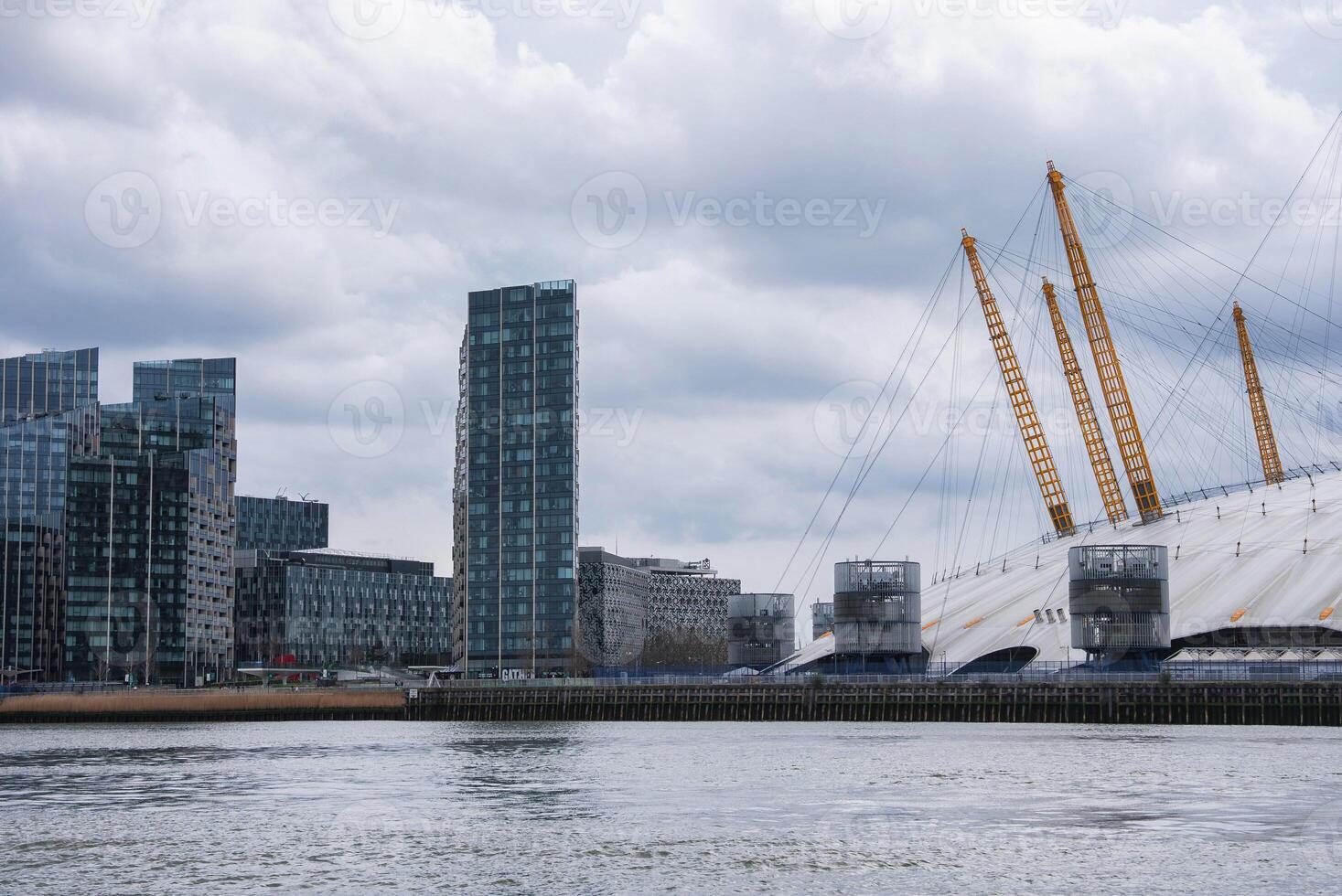 The O2 Arena's white dome and yellow masts stand out near modern high rise buildings by the River Thames under a cloudy London sky. photo