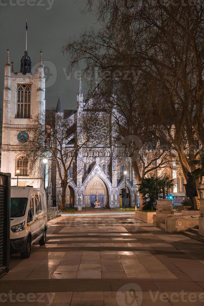 The lit facade of Westminster Abbey in London at night, framed by bare trees. A white van is parked on a paved walkway, with minimal activity visible. photo