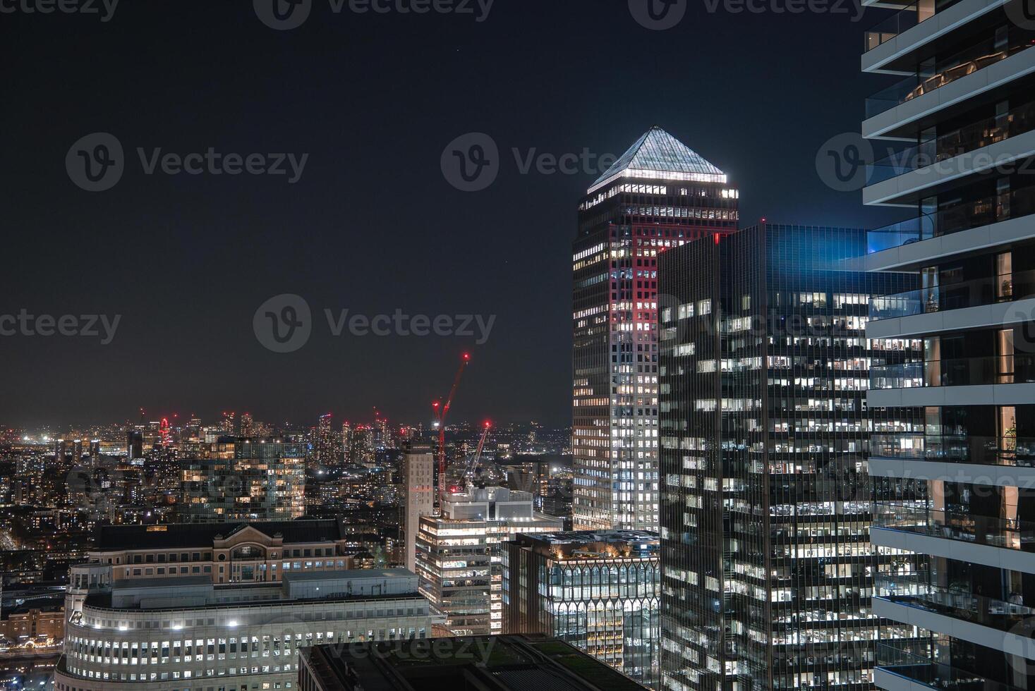 Illuminated skyscrapers of Canary Wharf, including One Canada Square, with glowing office lights, construction cranes, and a dark contrasting sky. photo