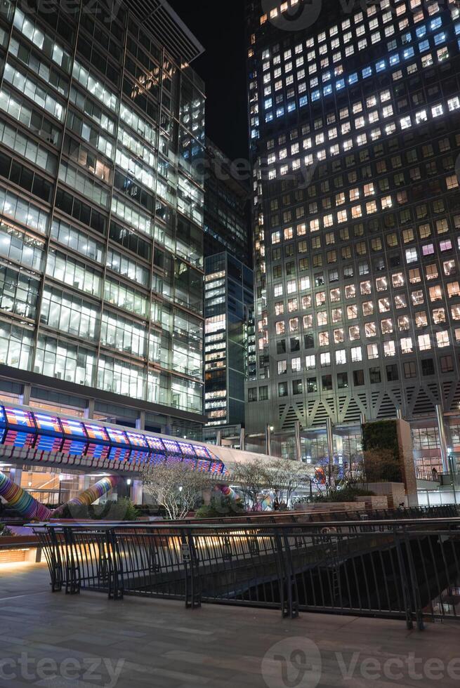 Canary Wharf at night with lit skyscrapers, a colorful tube like structure in the foreground, landscaped greenery, and urban design elements. photo