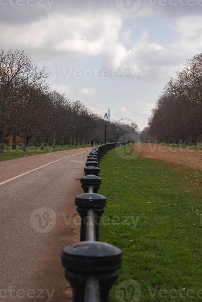 A serene park pathway in London with bare trees, black railings, and parallel paved and dirt paths. The London Eye is faintly visible in the distance. photo