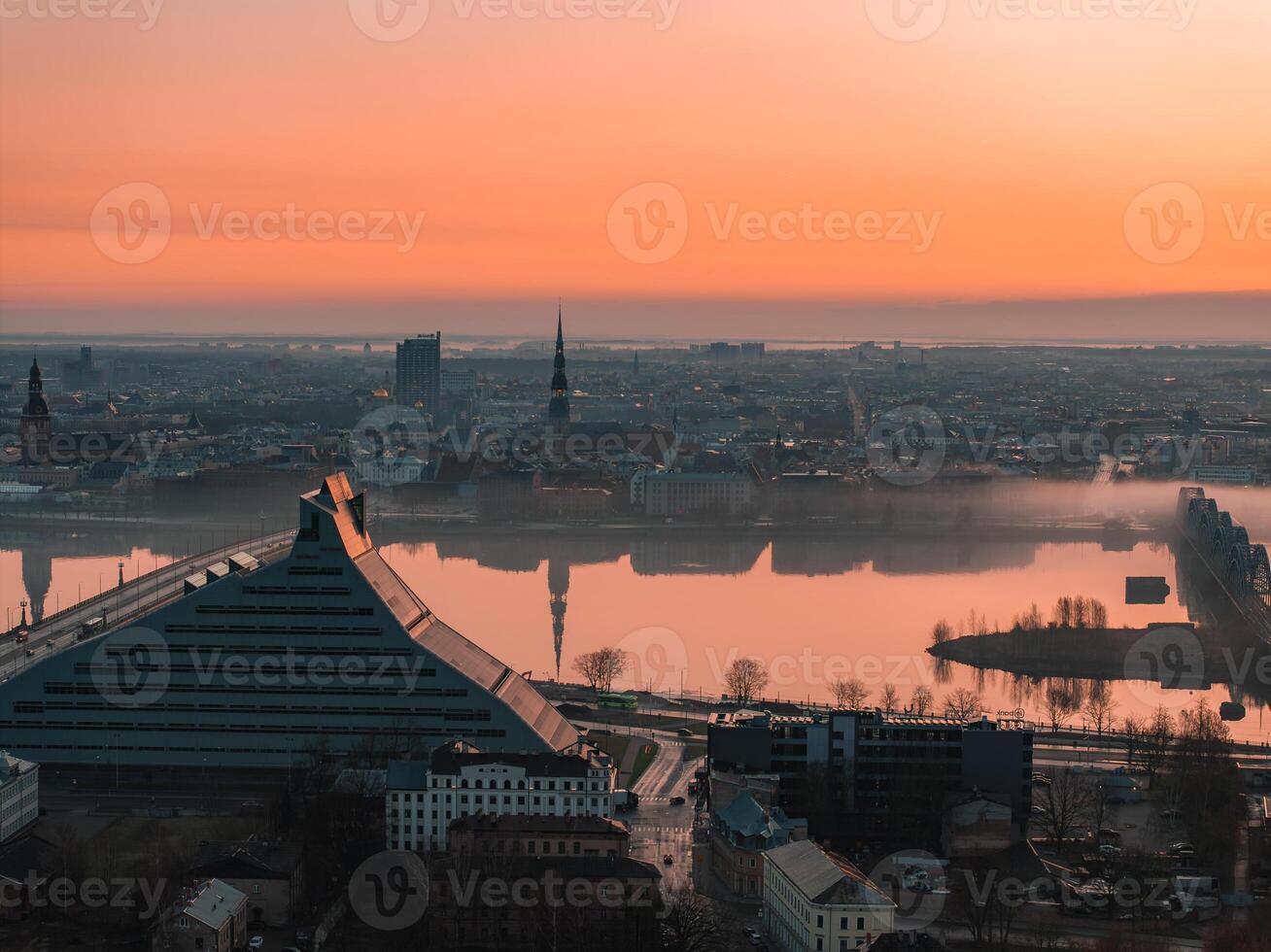 Serene sunrise view of Riga, Latvia, featuring the Daugava River, the National Library's angular design, and historic church spires in the background. photo