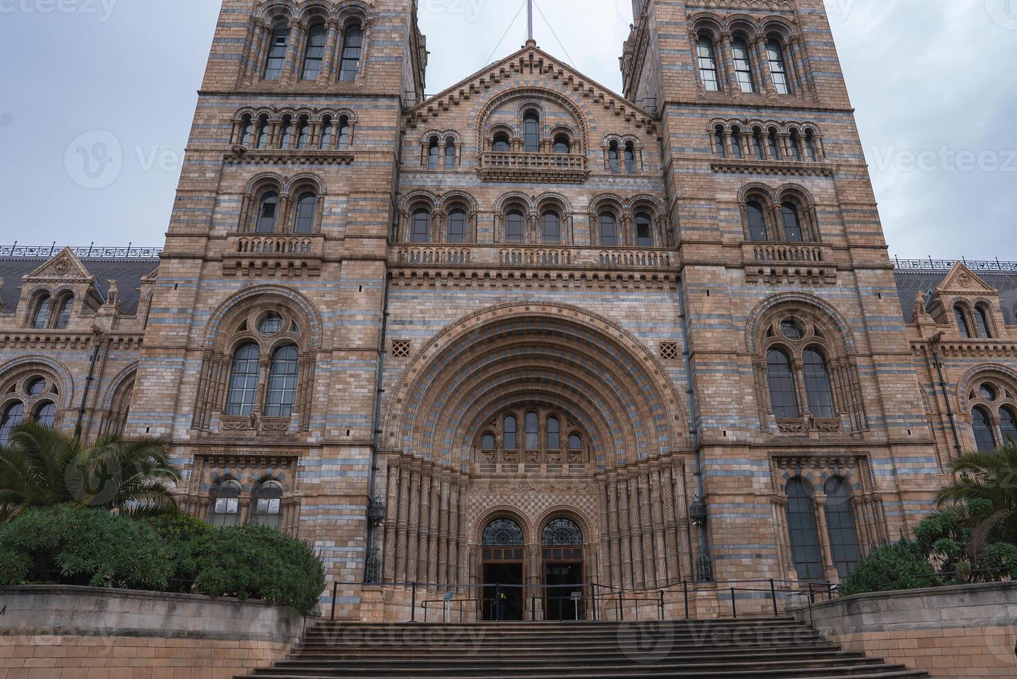 The grand facade of the Natural History Museum in London features Romanesque architecture, arched windows, tall towers, and greenery in the foreground. photo