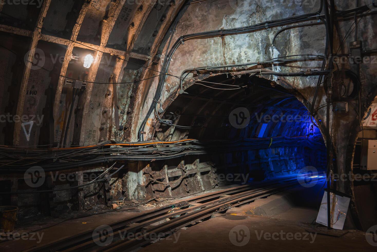 Aged underground tunnel in London with exposed concrete walls, structural reinforcements, and tracks leading into a dimly lit section with blue light. photo
