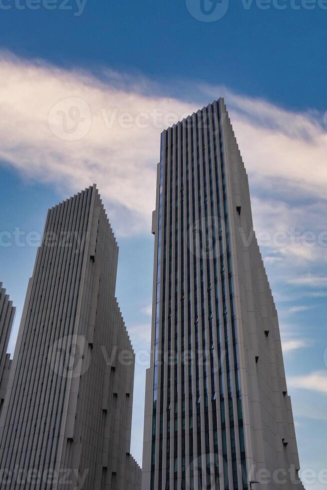 A group of sleek, vertical skyscrapers with white facades and narrow windows set against a clear blue sky with a streak of white clouds in London. photo