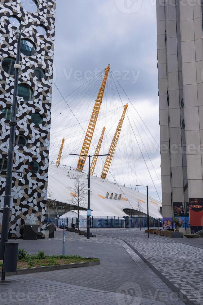 The O2 Arena's white dome and yellow masts stand out amid modern buildings, including one with a geometric facade, under a cloudy London sky. photo