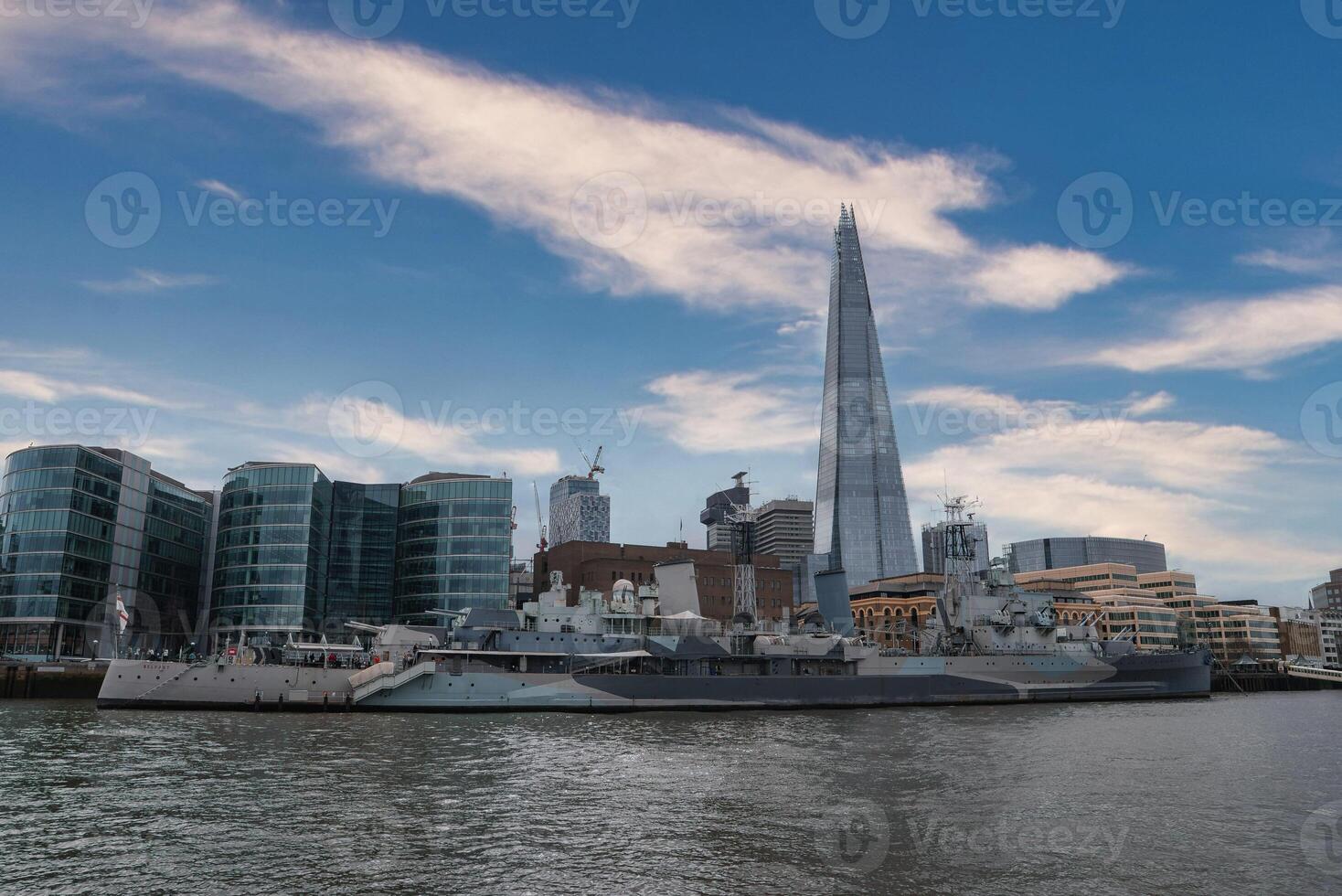 View of the Shard skyscraper towering over modern buildings, with HMS Belfast moored on the River Thames, showcasing London's mix of history and architecture. photo
