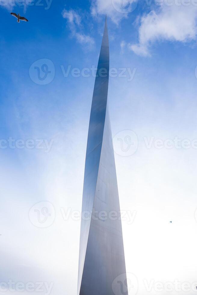 A sleek metallic structure with a pointed design rises against a bright blue sky with scattered clouds. A bird is in flight near the top left corner. photo