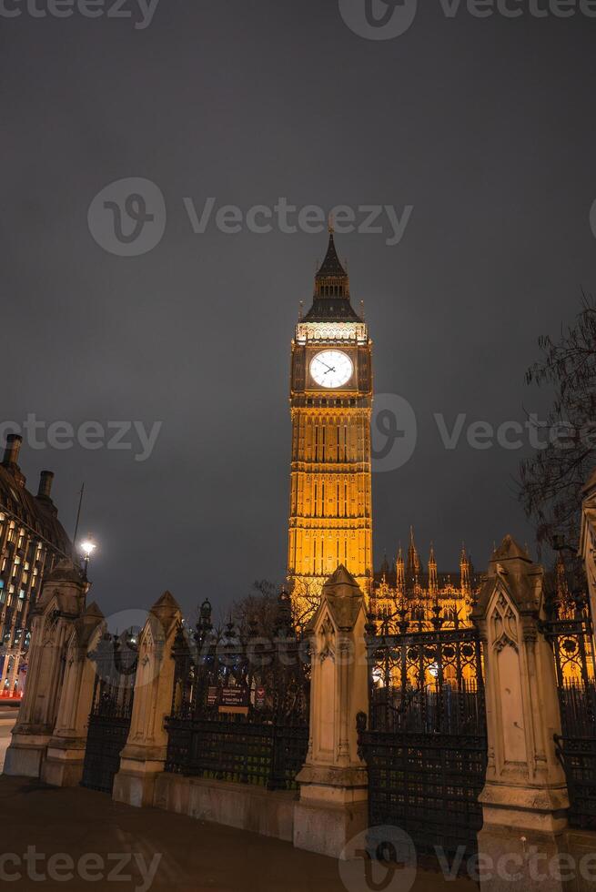 The iconic Big Ben clock tower, part of the Palace of Westminster, glows warmly at night. A wrought iron fence and nearby trees frame the scene. photo