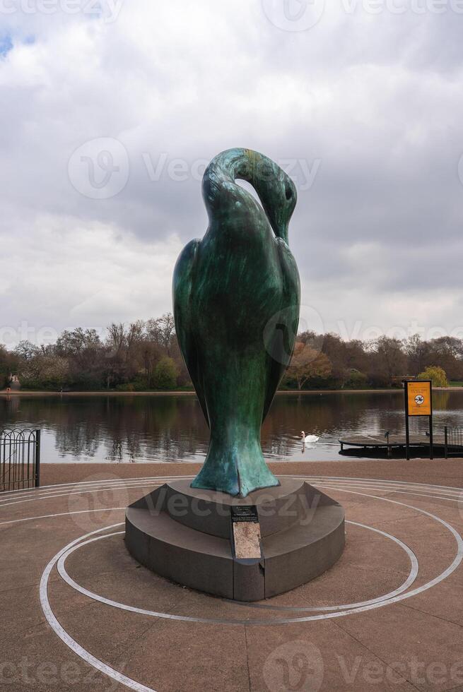 A green bronze bird sculpture on a circular pedestal near a calm lake with a swan, dock, autumn trees, and an overcast sky in a London park. photo
