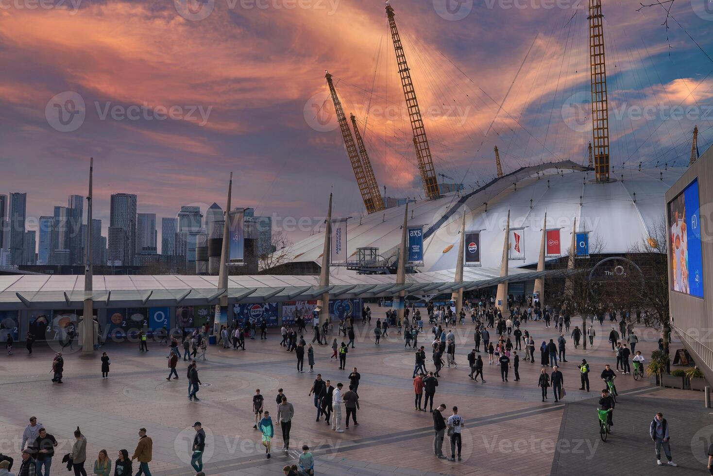 The O2 Arena's white dome and yellow towers stand out in London, with a busy plaza in the foreground and Canary Wharf's skyline in the background. photo
