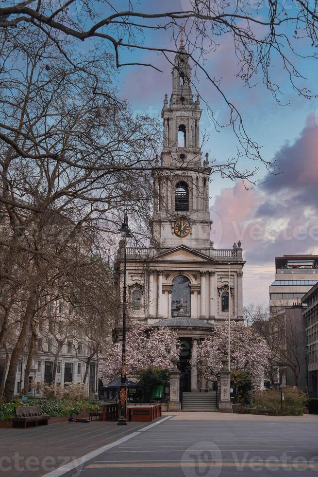 Baroque style St. Mary le Strand church in London, surrounded by trees in bloom, with a paved area, benches, and modern buildings in the background. photo