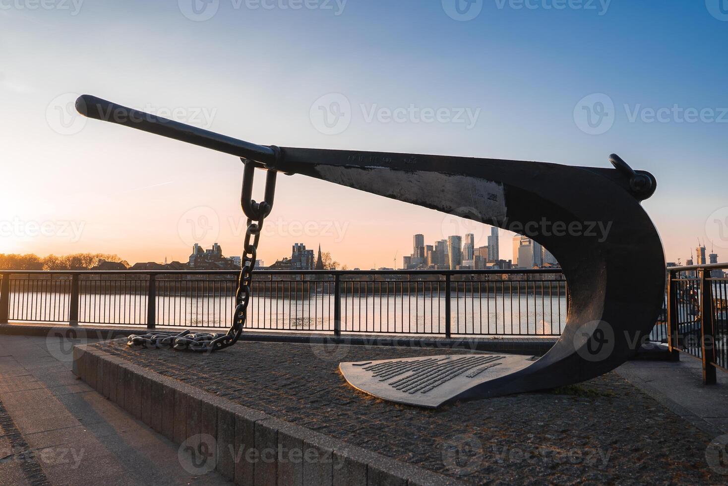 A large black anchor sculpture on a raised platform near the Thames River, with Canary Wharf's modern skyscrapers glowing in a warm sunset light. photo