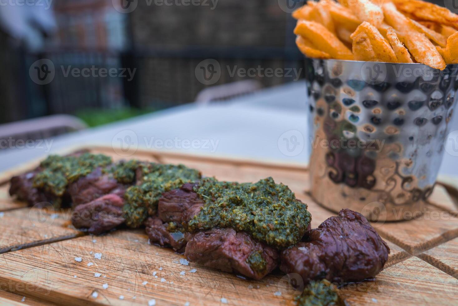 Close up of grilled steak slices with green herb sauce on a wooden board, paired with fries in a metal cup. Outdoor dining with brick and greenery in London. photo
