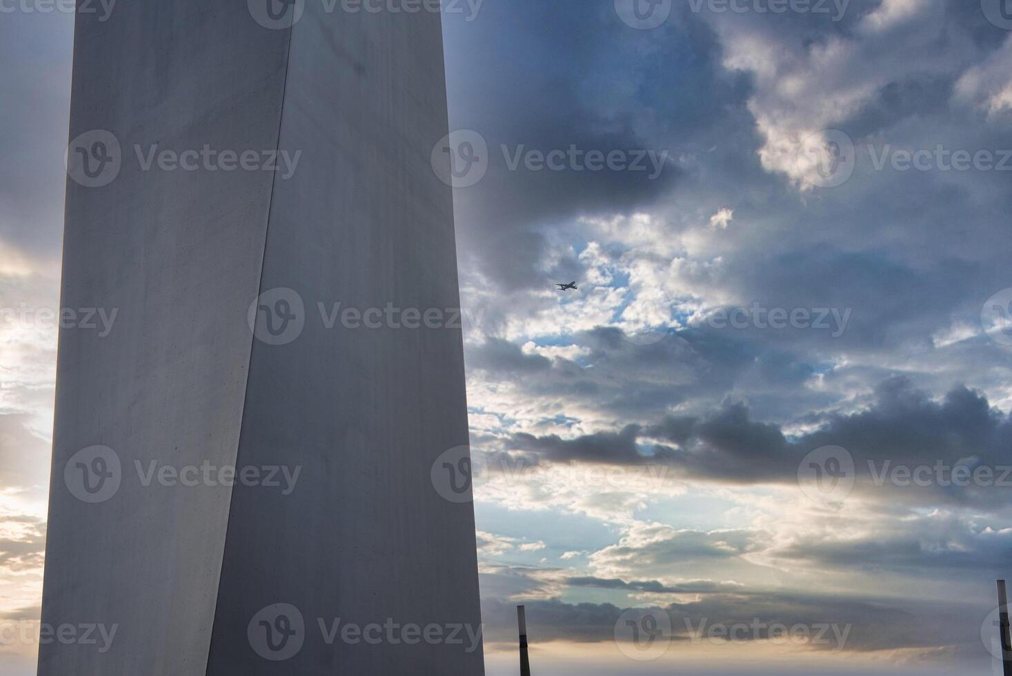 A tall, angular structure with a smooth surface stands in the foreground. A small airplane flies in a partly cloudy sky illuminated by soft sunlight. photo