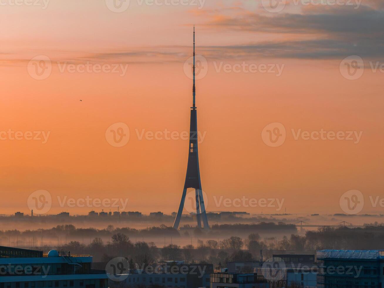 Serene morning scene in Riga, Latvia, featuring the iconic Riga TV Tower with its tripod design, light mist, and city rooftops under an orange sunrise. photo