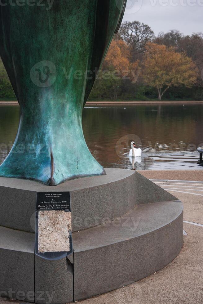 A green patinated sculpture on a stone pedestal with a plaque near a calm body of water, a white swan, and autumn colored trees in a London park. photo