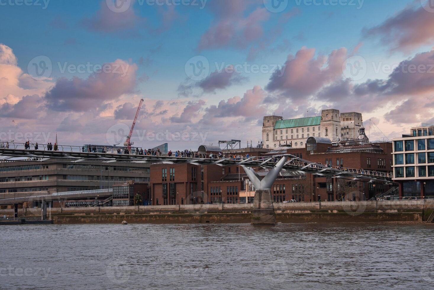 Millennium Bridge in London spans the River Thames, busy with pedestrians. Tate Modern and surrounding architecture are visible under a pink and blue sky. photo