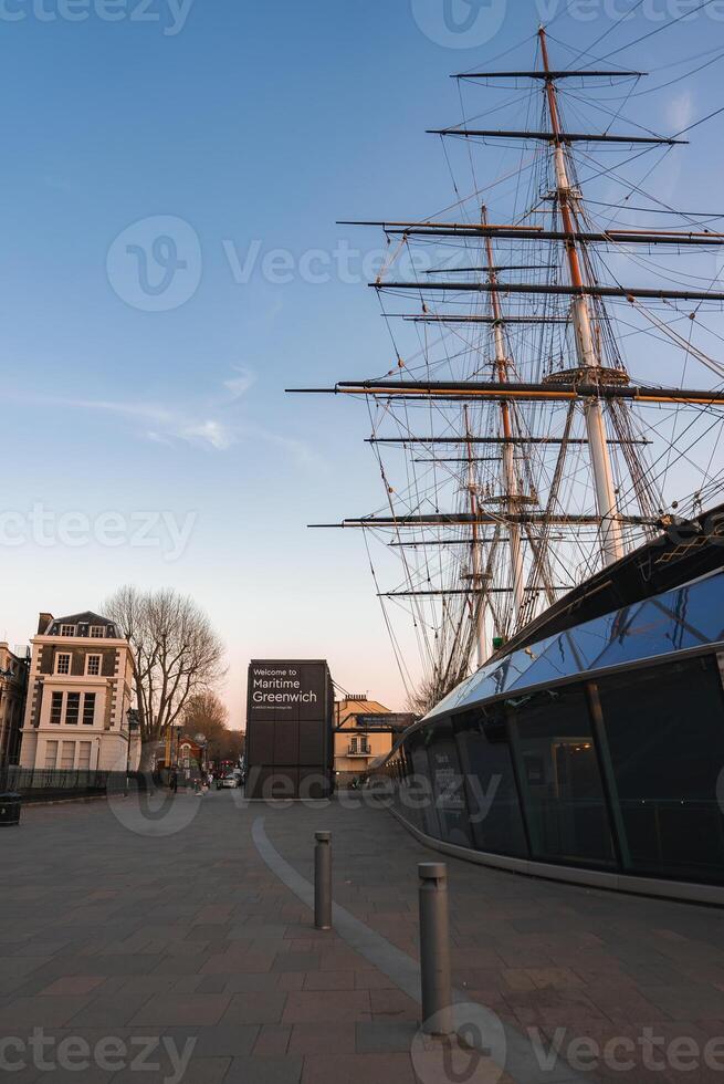 The Cutty Sark's tall masts and rigging stand above a modern glass base in Greenwich, London, with a Maritime Greenwich sign and nearby trees. photo