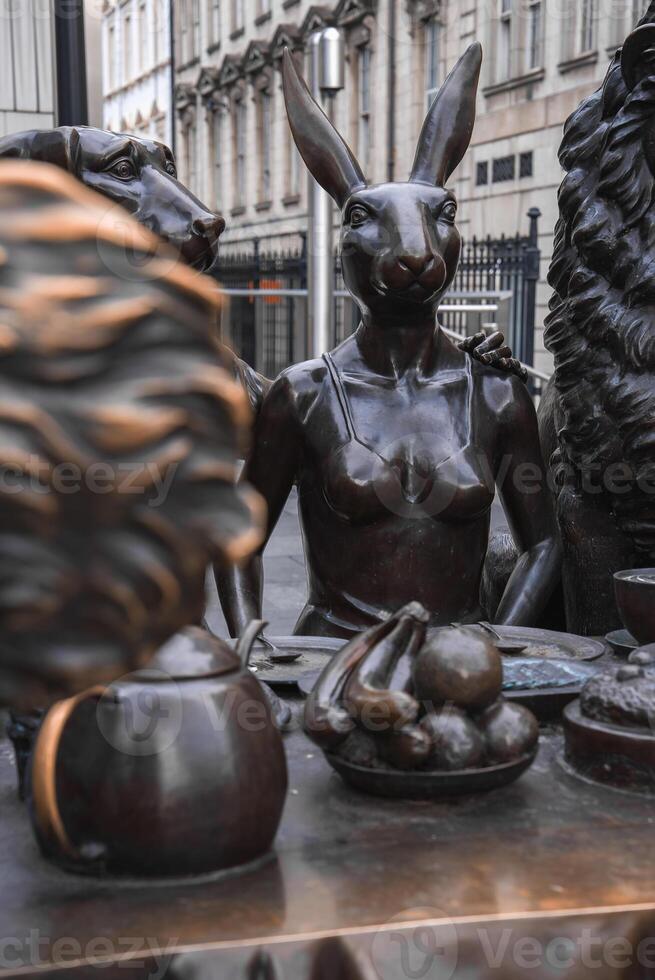 A bronze sculpture of a rabbit, dog, and lion around a table with a teapot and fruit, set against historic style buildings and black iron fencing in London. photo