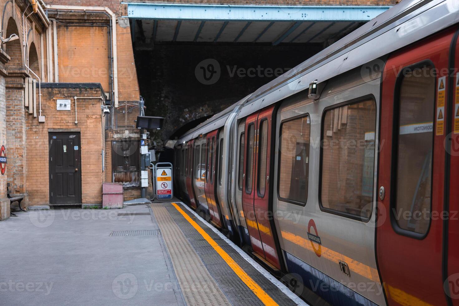 A London Underground train stands at a station platform with brick walls, arched details, a yellow safety line, and a visible tunnel entrance in the background. photo