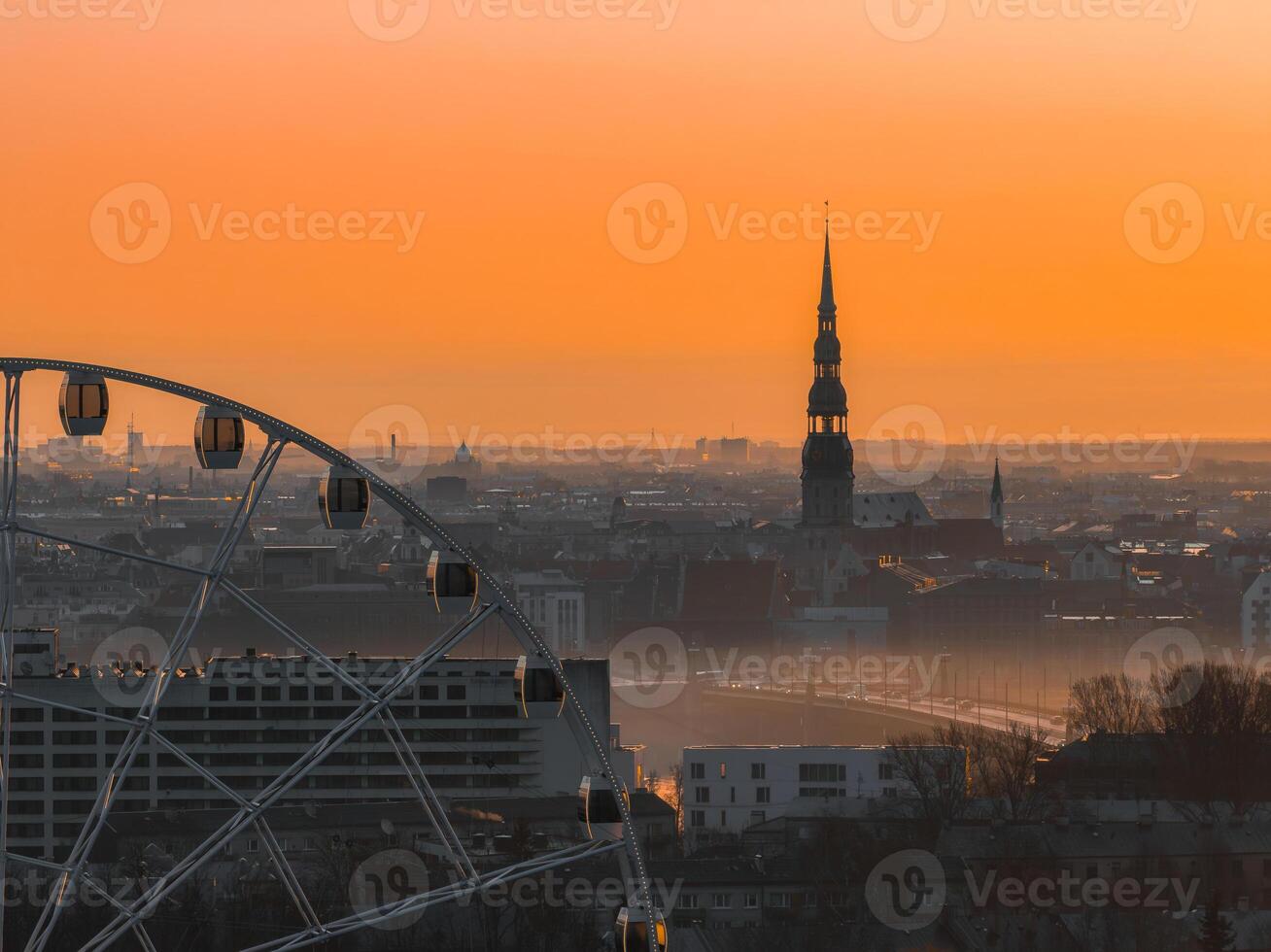 Serene sunrise over Riga, Latvia, featuring St. Peter's Church spire, a Ferris wheel in the foreground, and a warm orange sky with soft morning light. photo